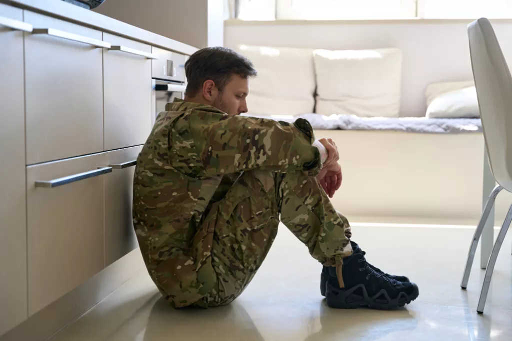 Upset soldier sits on floor hugging his knees, in sunny room, he looks at floor, he wearing field military clothes