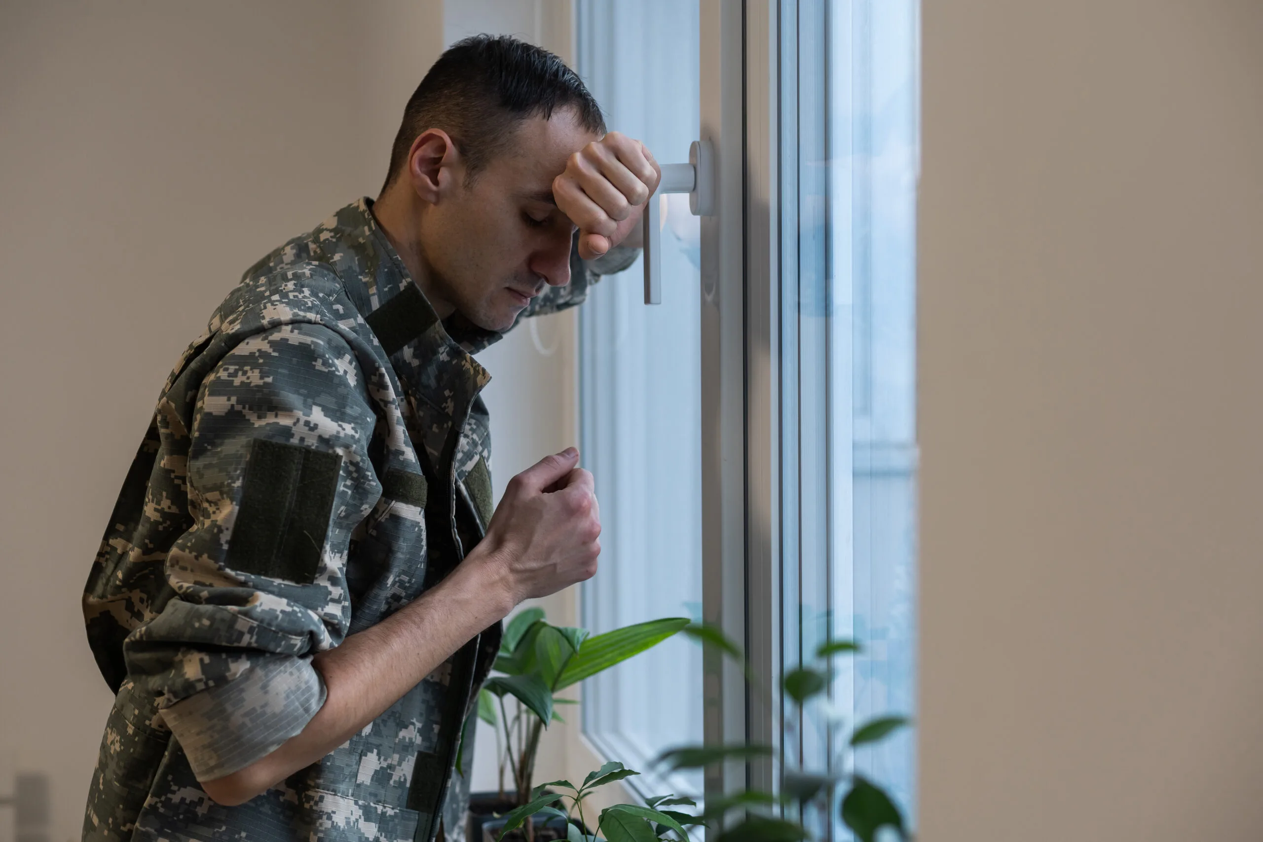 Depressed and sad soldier in green uniform with trauma after war standing near the window.