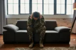 Full length shot of middle aged military man holding his head in pain and depression sitting on the couch. Soldier suffering from psychological trauma. PTSD concept. Front view. Horizontal shot