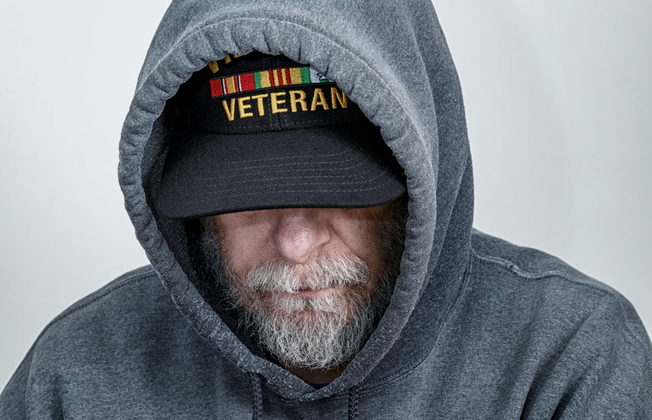 A Vietnam War USA military veteran is looking down with his partially obscured face showing a serious facial expression. Though this shot is posed, this is a real life, real person Vietnam war veteran who's recently had some significant health issues. He is wearing an inexpensive, non-branded, generic, souvenir shop replica Vietnam veteran commemorative baseball hat style cap under his hoody sweatshirt hood.